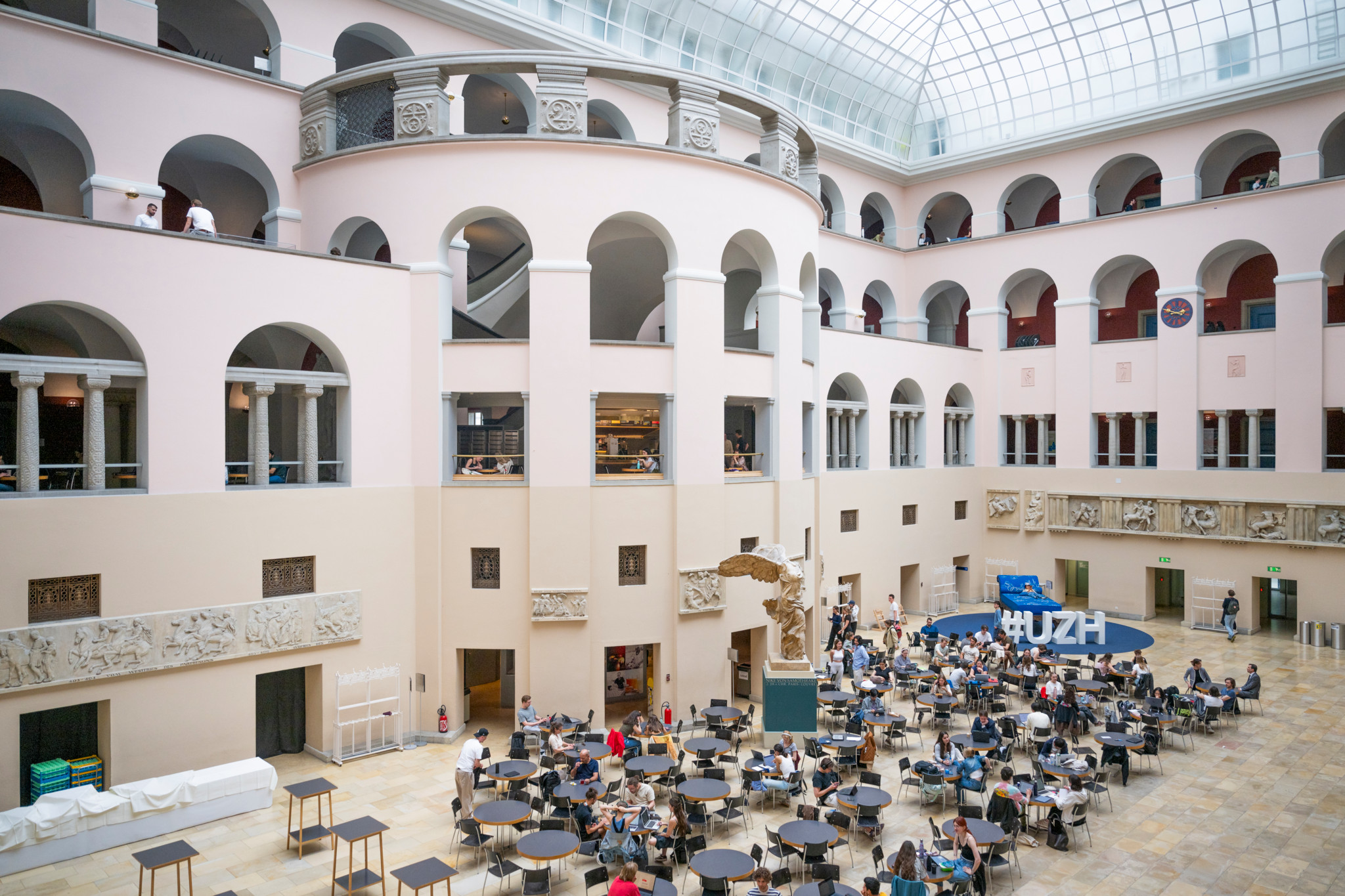 Lichthof der Universität Zürich mit Studierenden an Tischen unter dem Glasdach, mehrere Stockwerke mit Rundbögen und UZH-Banner sichtbar.