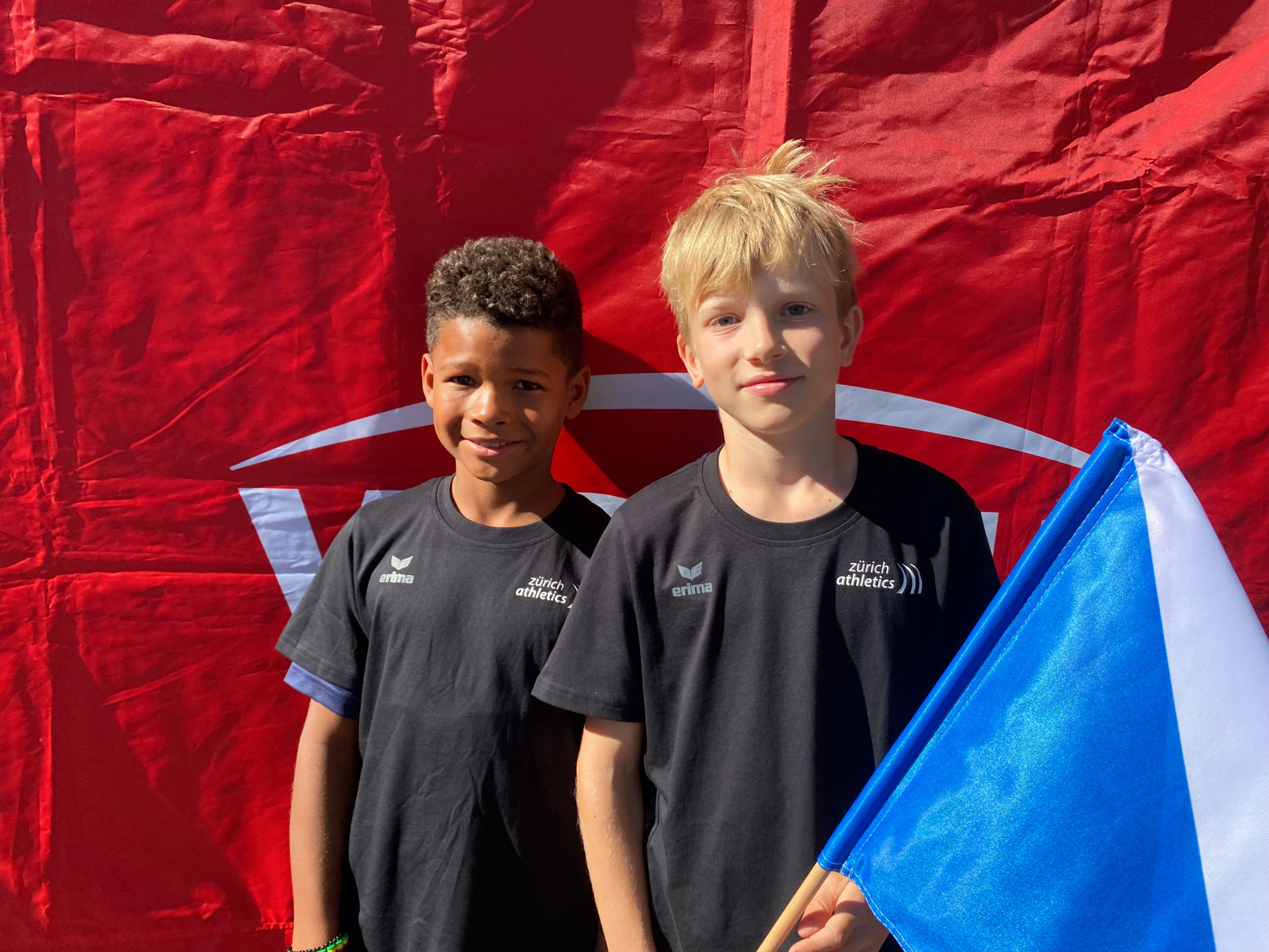 Zwei junge Jungen in schwarzen T-Shirts vor einem roten Hintergrund, einer hält eine blaue und weisse Flagge.