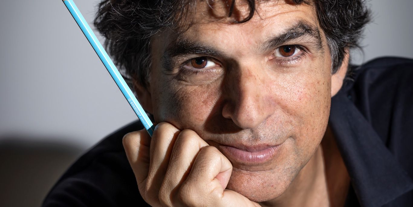 Homme aux cheveux noirs bouclés portant une chemise sombre, tenant un crayon bleu près de son visage.