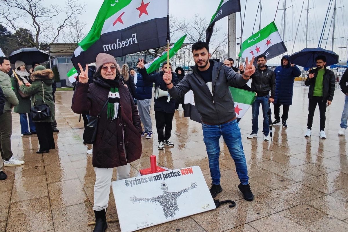 Manifestation sur une place: Wajd Zimmermann du collectif Femmes syriennes pour la démocratie à gauche avec des drapeaux syriens et pancarte.