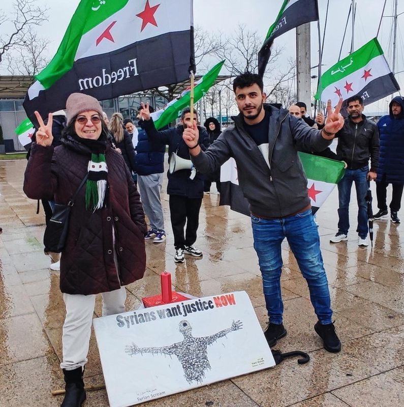 Manifestation sur une place: Wajd Zimmermann du collectif Femmes syriennes pour la démocratie à gauche avec des drapeaux syriens et pancarte.