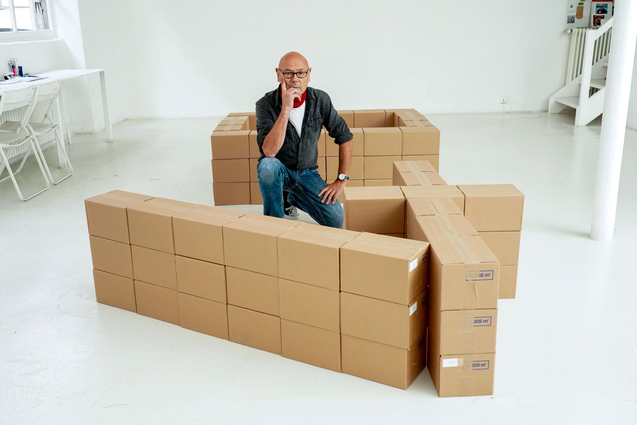 Un homme chauve portant des lunettes rouges, accroupi au milieu d’une structure formée de nombreux cartons en carton brun dans un espace blanc. Un homme chauve portant des lunettes rouges, accroupi au milieu d’une structure formée de nombreux cartons en carton brun dans un espace blanc.