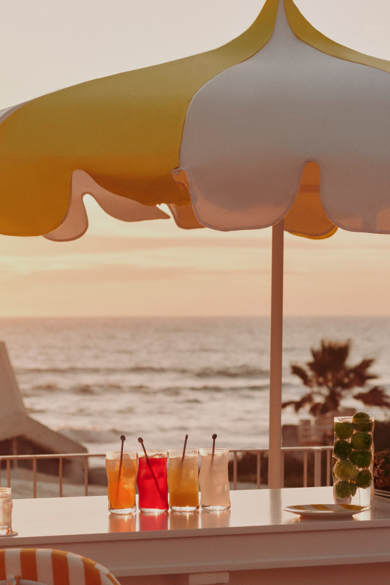 Bunte Cocktails auf einem Tisch unter einem gelb-weissen Sonnenschirm, mit Meerblick und Sonnenuntergang im Hintergrund.