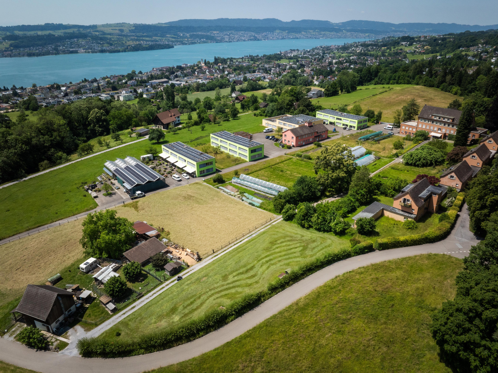 Luftaufnahme eines Schulcampus mit Gewächshäusern, Grünflächen und einem See im Hintergrund, umgeben von Hügeln und Wohngebäuden.