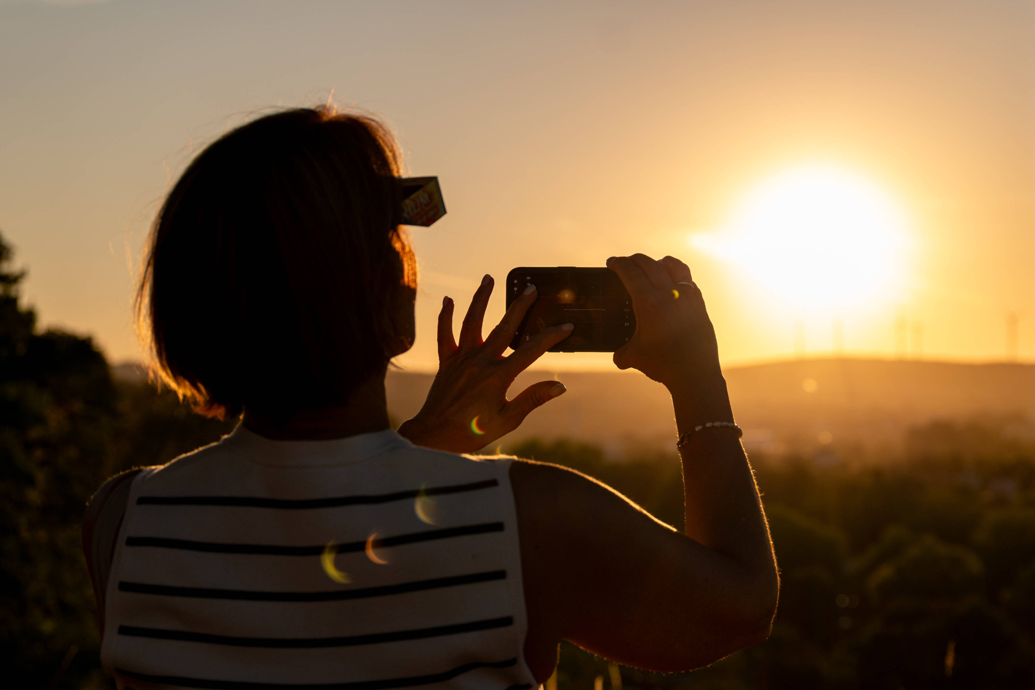 Frau mit Sonnenfinsternisbrille fotografiert die partielle Sonnenfinsternis vom 12. August 2026 mit ihrem Smartphone vor tiefstehender Sonne.