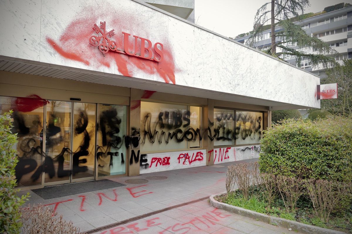Façade d’une banque UBS vandalisée avec des graffitis en rouge et noir, incluant des inscriptions telles que ’Palestine’.