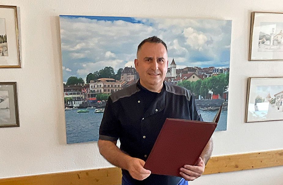 Un chef de restaurant tenant un menu, se tenant devant un tableau représentant un paysage urbain.