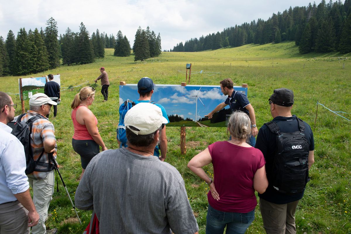 Un groupe de personnes écoute des explications près d'un panneau d'affichage avec des photomontages sur le parc éolien de la Grandsonnaz, avec des paysages verdoyants en arrière-plan.