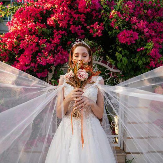Mariée en robe blanche tenant un bouquet de fleurs roses devant un buisson de bougainvilliers en fleurs.
