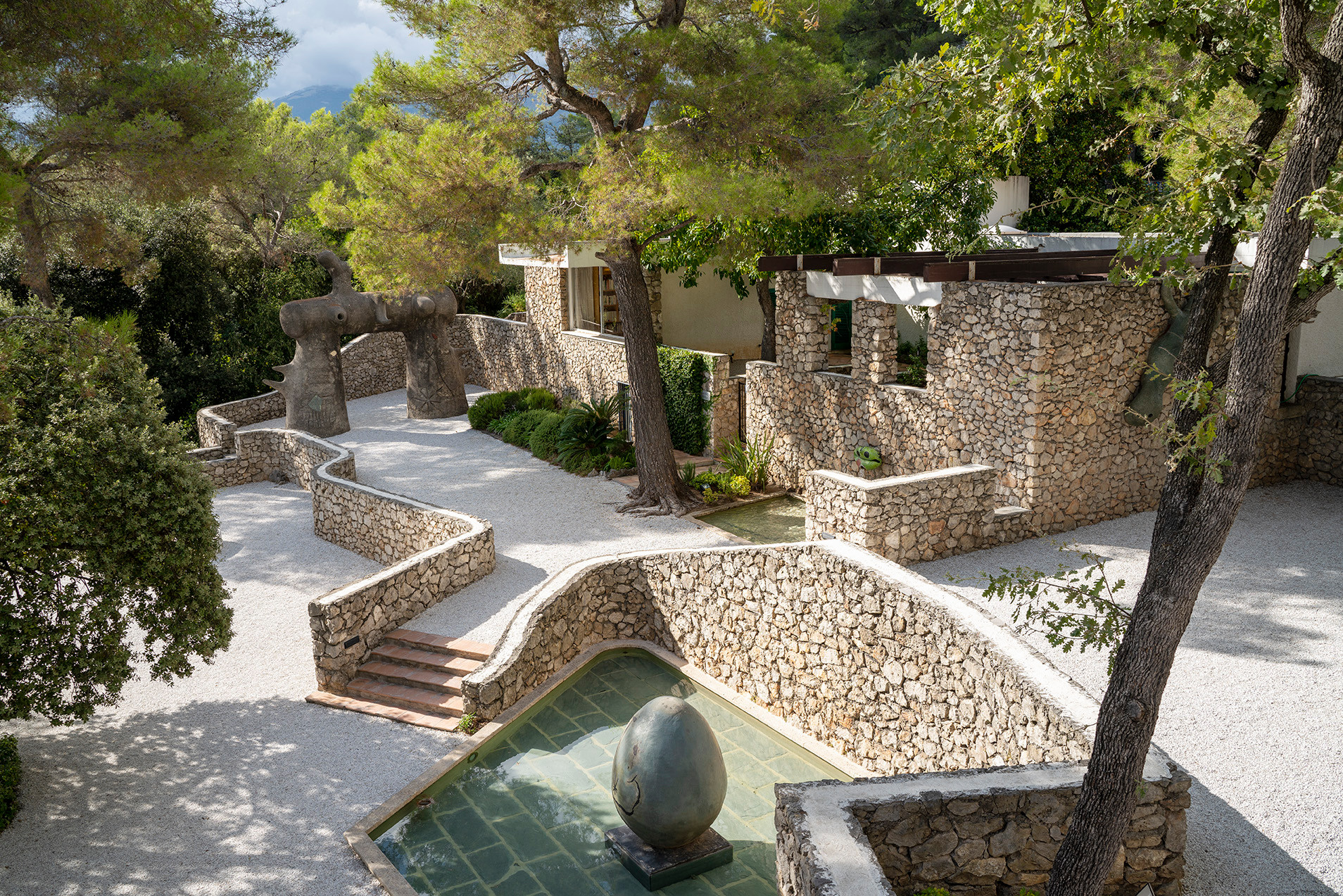 Mediterraner Garten mit Natursteinmauern, geschwungenen Treppen, einem Wasserbecken mit Steinkugel und alten Bäumen.