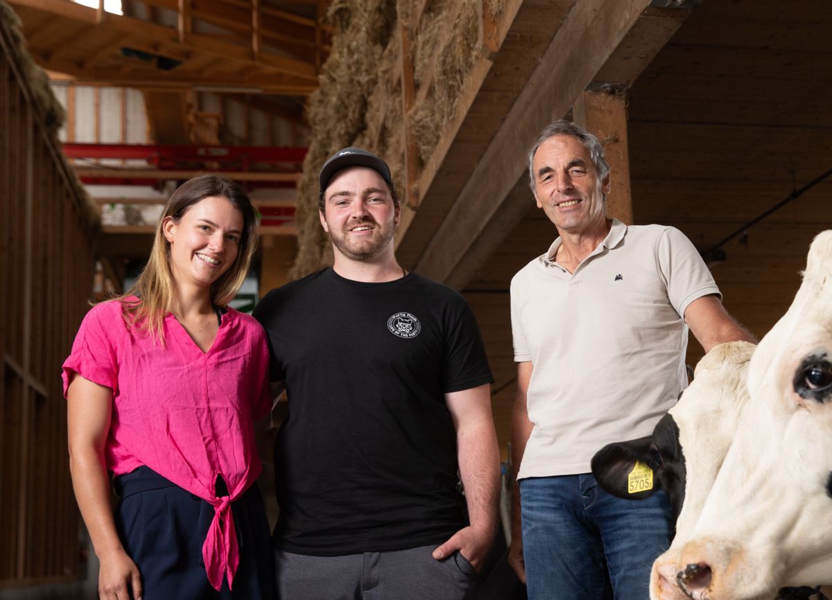 Drei Personen stehen in einem Stall neben einer Kuh mit schwarz-weissen Flecken.