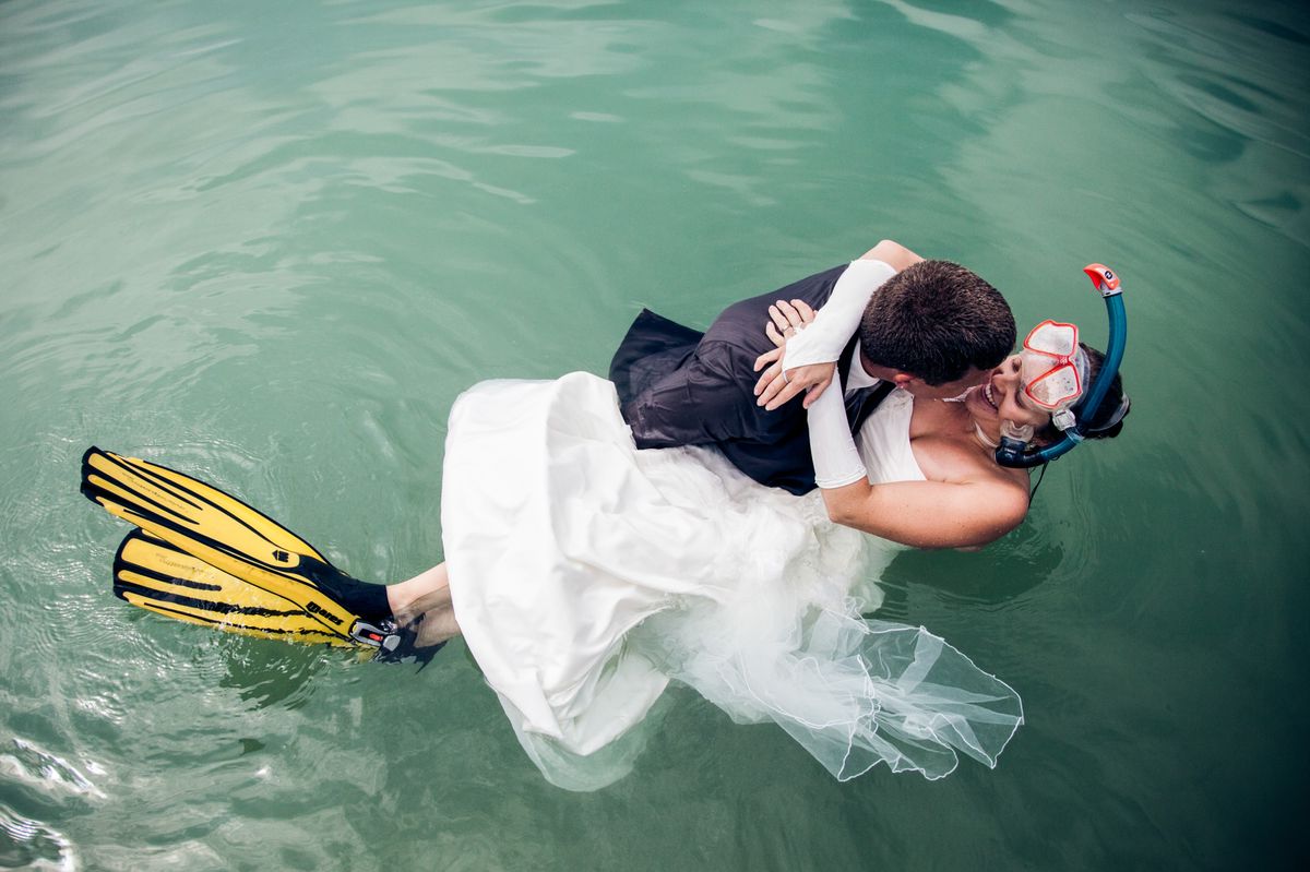 Un couple en tenue de mariage, avec la mariée portant des palmes et un masque de plongée, s'embrasse dans l'eau.