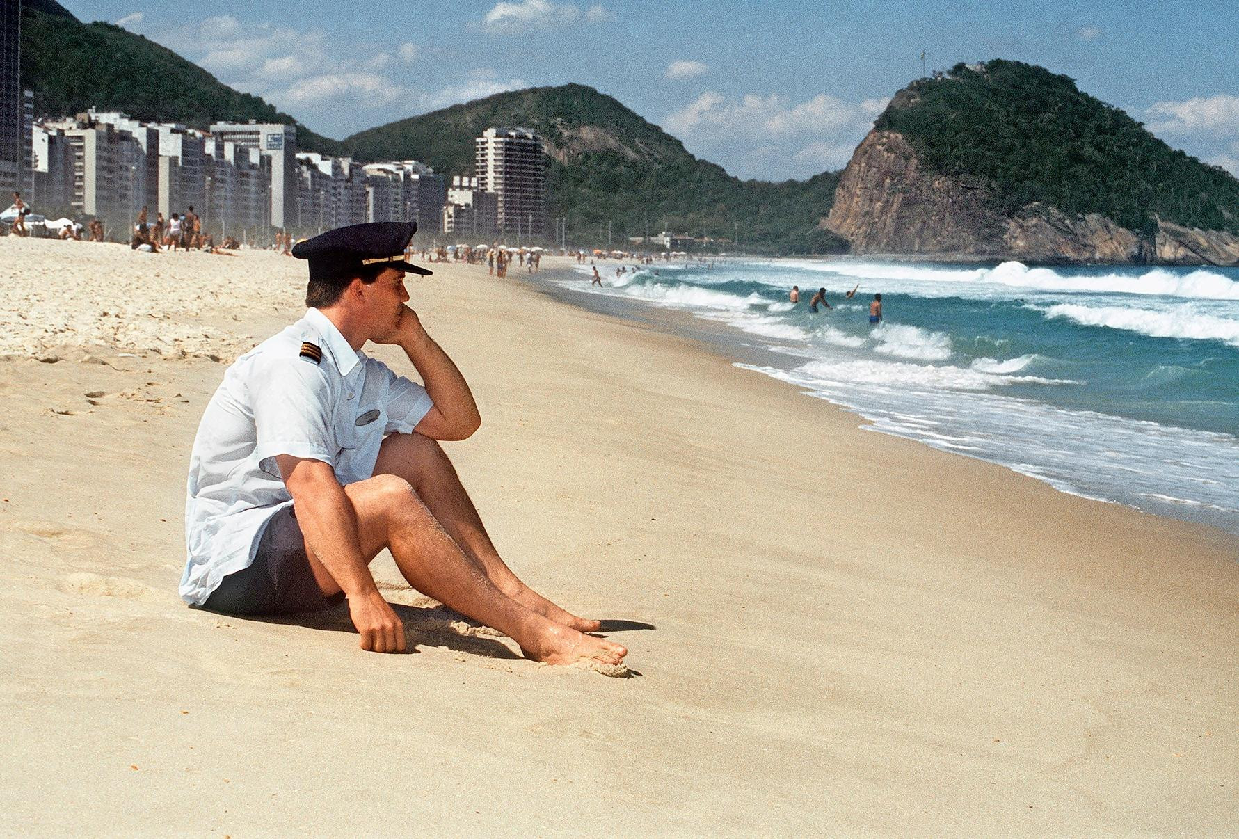 Mann in Uniform mit Schirmmütze sitzt barfuss nachdenklich am Sandstrand von Rio de Janeiro mit Stadtsilhouette und Bergen im Hintergrund.