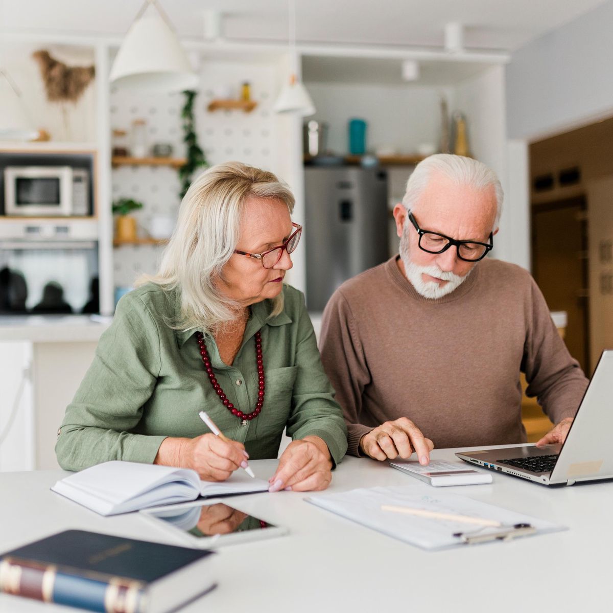 Un couple senior examine ses finances à domicile en utilisant un ordinateur portable, des cahiers posés sur la table.