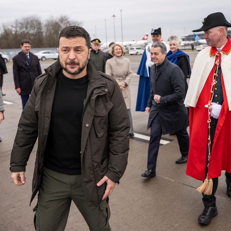 Un groupe de personnes marchant sur le tarmac d’un aéroport, avec un homme en manteau rouge et chapeau noir en avant-plan.