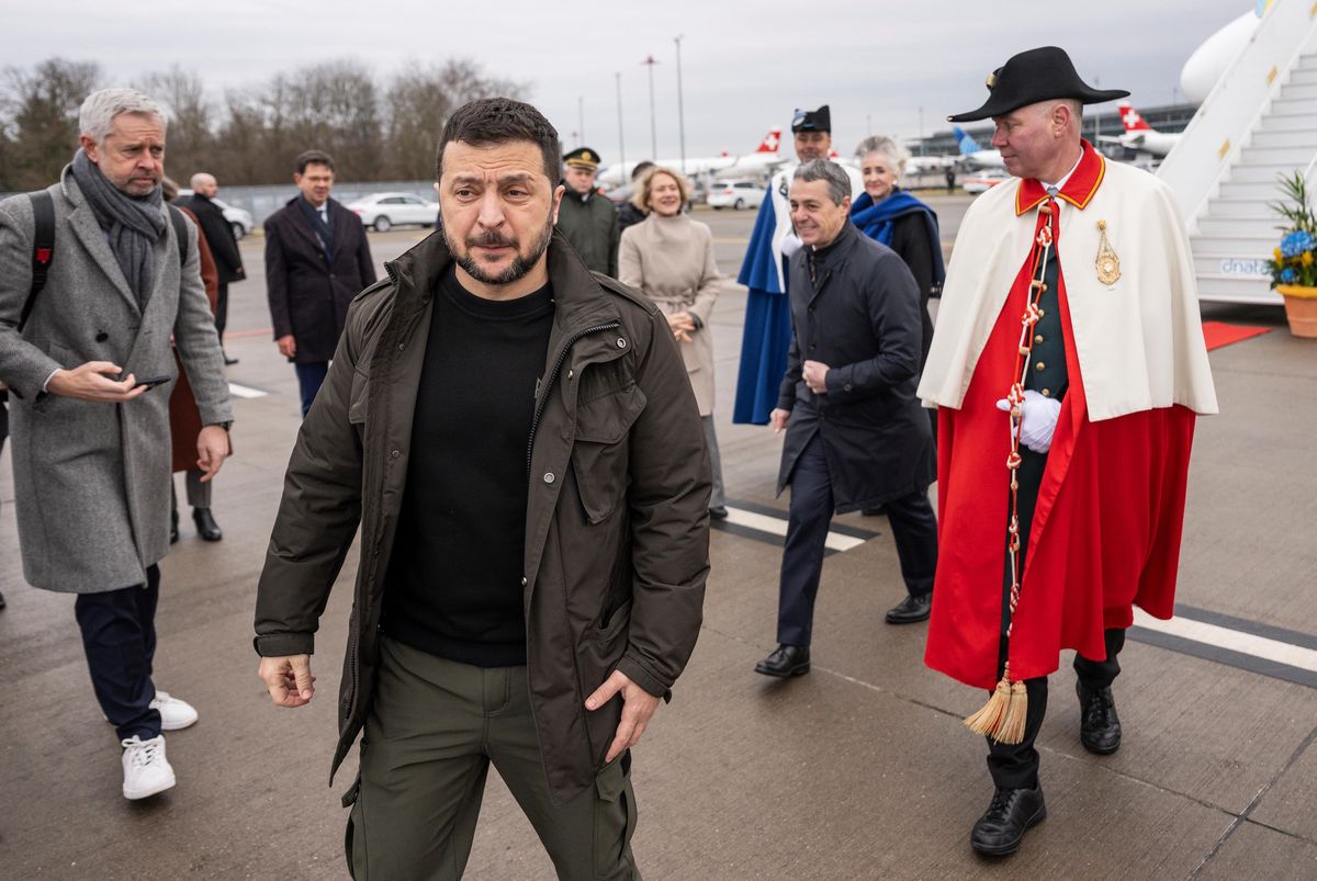 Un groupe de personnes marchant sur le tarmac d’un aéroport, avec un homme en manteau rouge et chapeau noir en avant-plan.