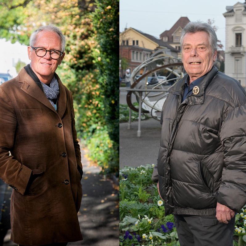 Zwei ältere Männer in einem herbstlichen Aussenbereich; links ein Mann in braunem Mantel und Schal, rechts ein Mann in grauer Daunenjacke vor einem Brunnen.