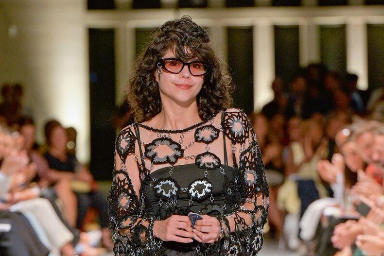 Femme portant une robe noire élégante et des lunettes de soleil lors d’un défilé de mode, entourée d’un public attentif.