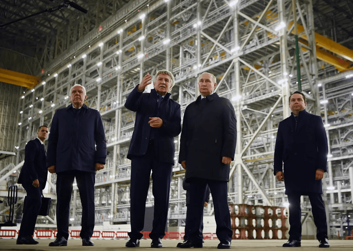 Groupe de cinq hommes en costume sombre dans un grand hangar industriel avec une structure métallique imposante en arrière-plan.