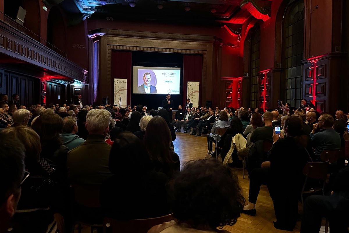 Un événement se déroule dans une grande salle illuminée par des lumières rouges, avec un écran projetant une présentation à un public nombreux.