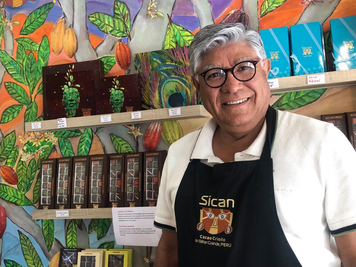 Un homme souriant portant un tablier noir 'Sican' se tient devant des étagères remplies de boîtes de chocolat colorées, avec un mur vibrant de motifs végétaux en arrière-plan.