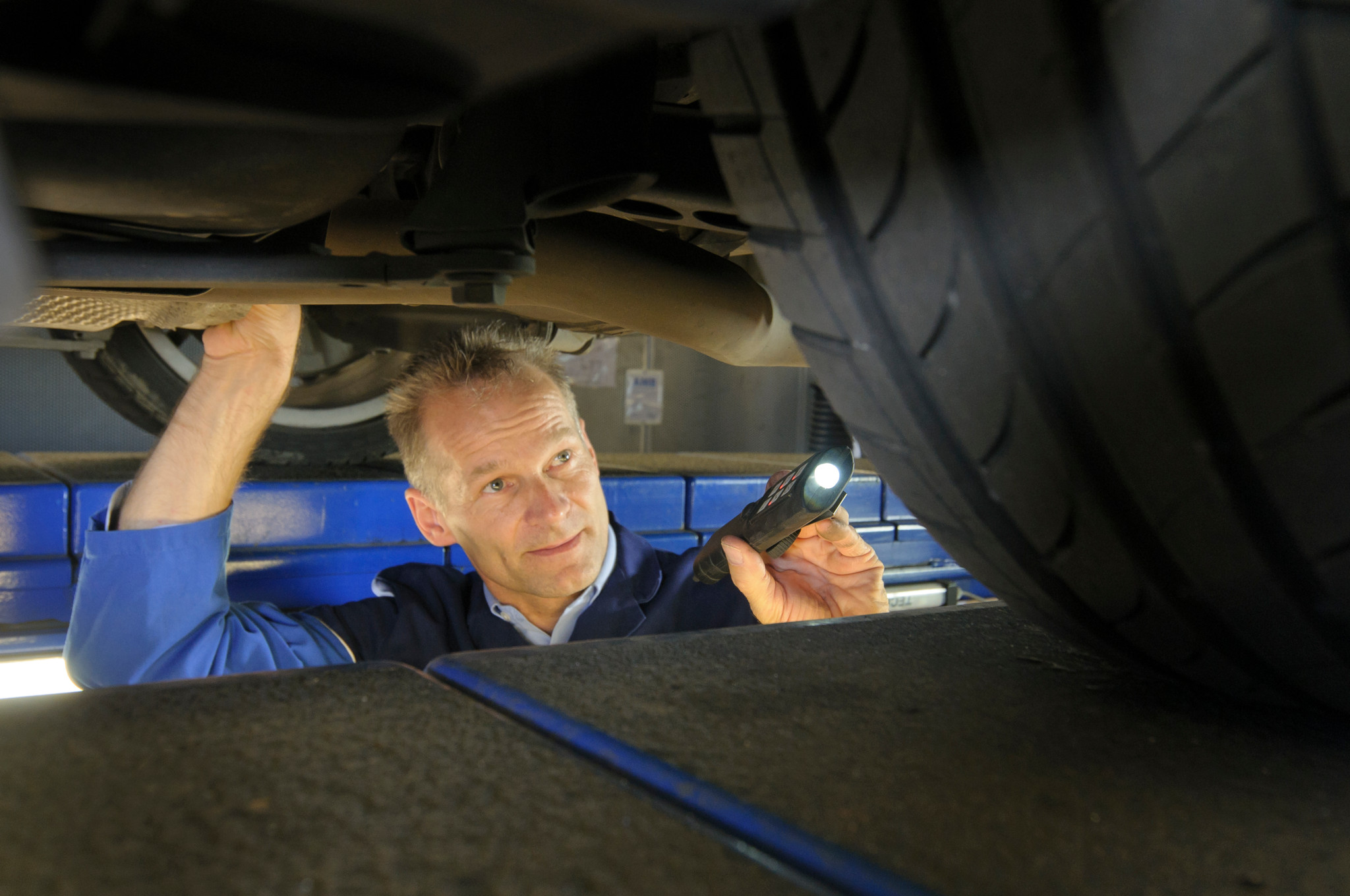 TÜV Rheinland Proficheck: Mechaniker leuchtet mit Taschenlampe unter ein Auto und prüft sicherheitsrelevante Teile für Fahranfänger.