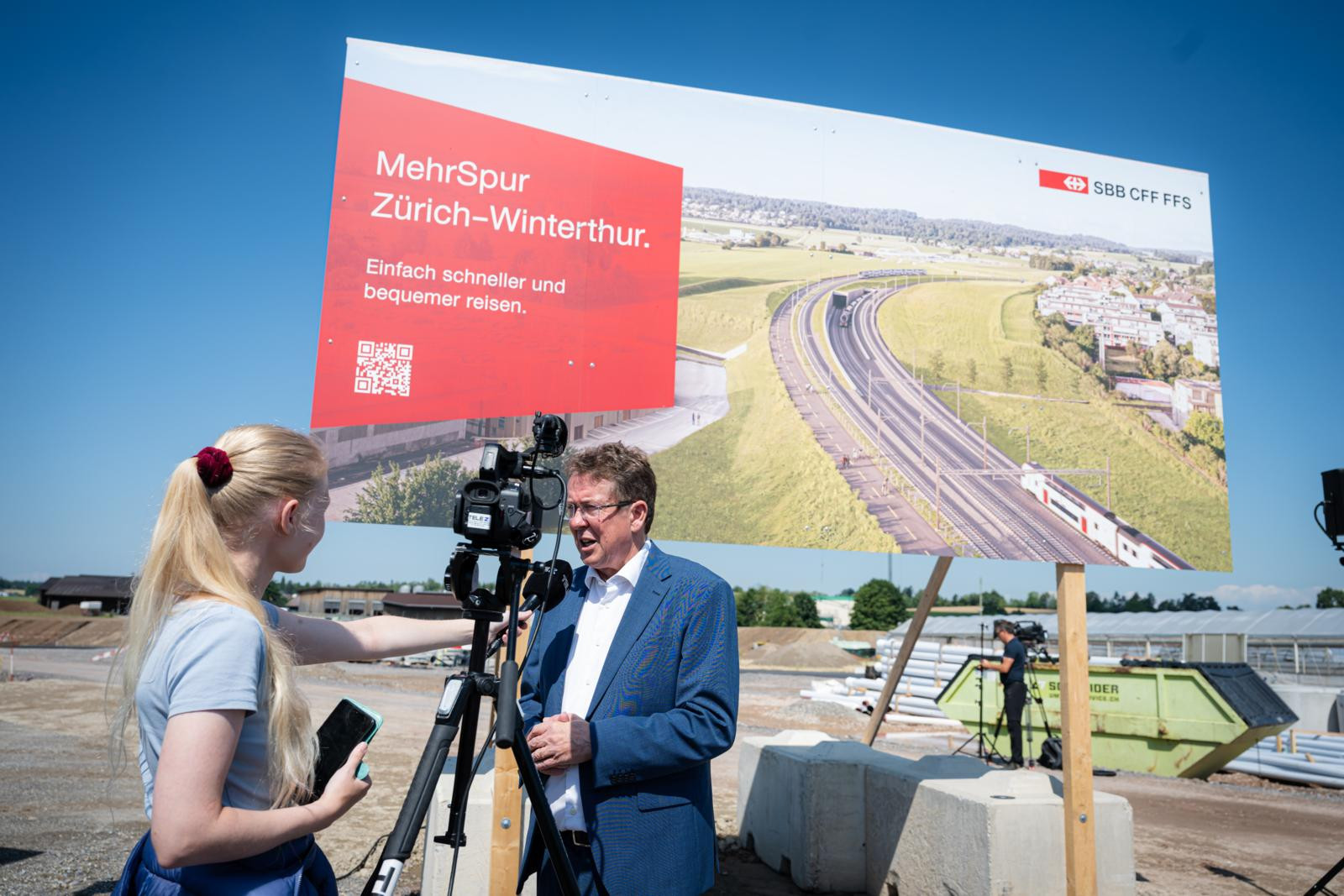 Mann gibt Interview vor SBB-Plakat «MehrSpur Zürich–Winterthur. Einfach schneller und bequemer reisen.» bei einer Baustelle.