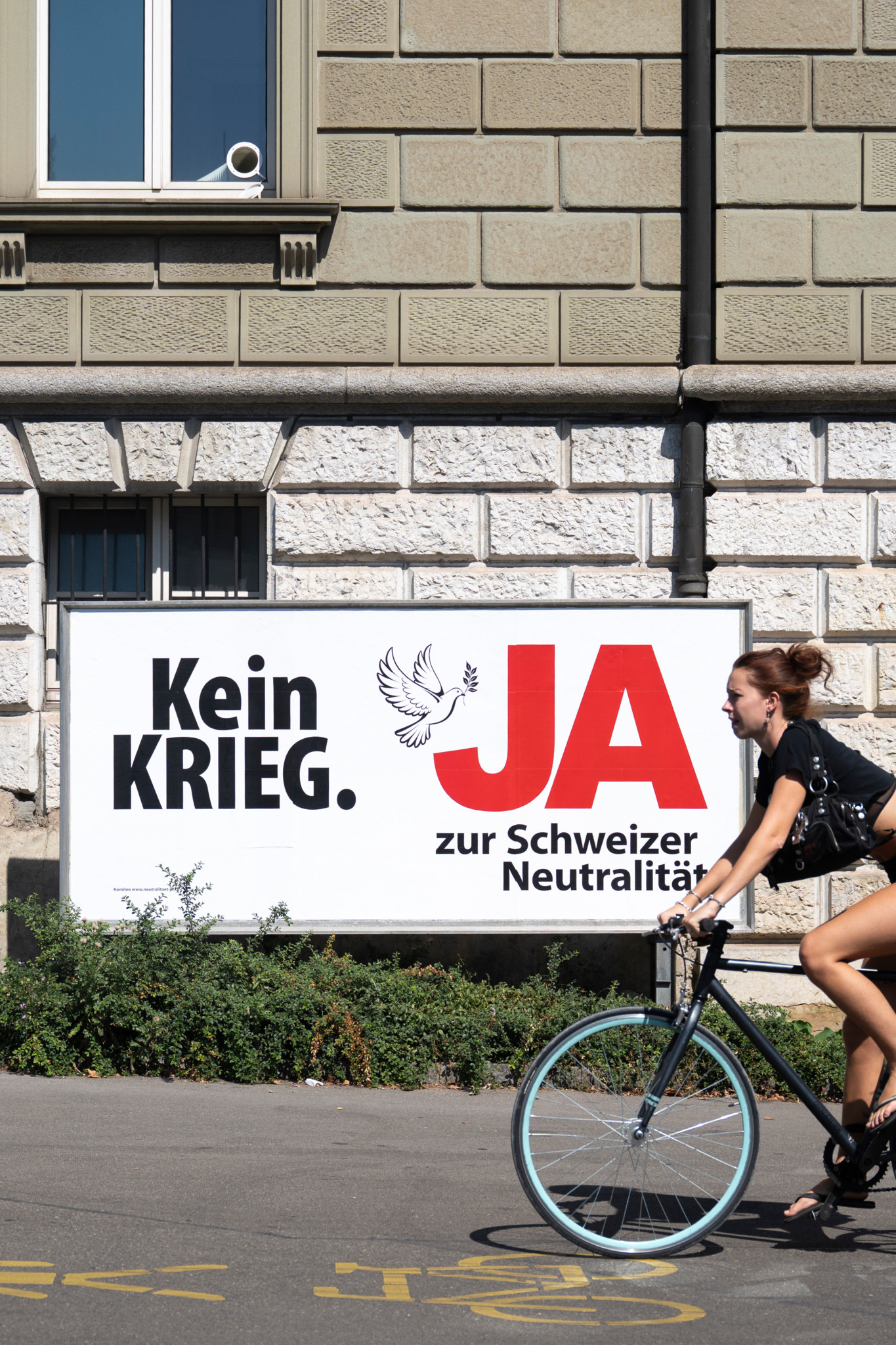 Frau auf Fahrrad fährt an Plakat zur Neutralitäts-Initiative vorbei: «Kein Krieg. JA zur Schweizer Neutralität»