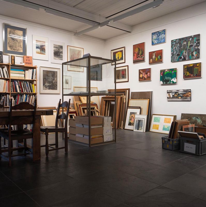 Intérieur de la galerie Roch à Ballens avec des œuvres d’art accrochées aux murs, une table et des chaises en bois, et des livres sur une étagère.