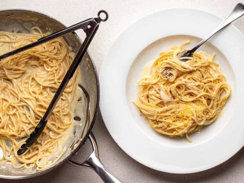 Cremige Spaghetti in einer Pfanne mit Zange, daneben ein weisser Teller mit Pasta und geriebenem Käse, von oben fotografiert.