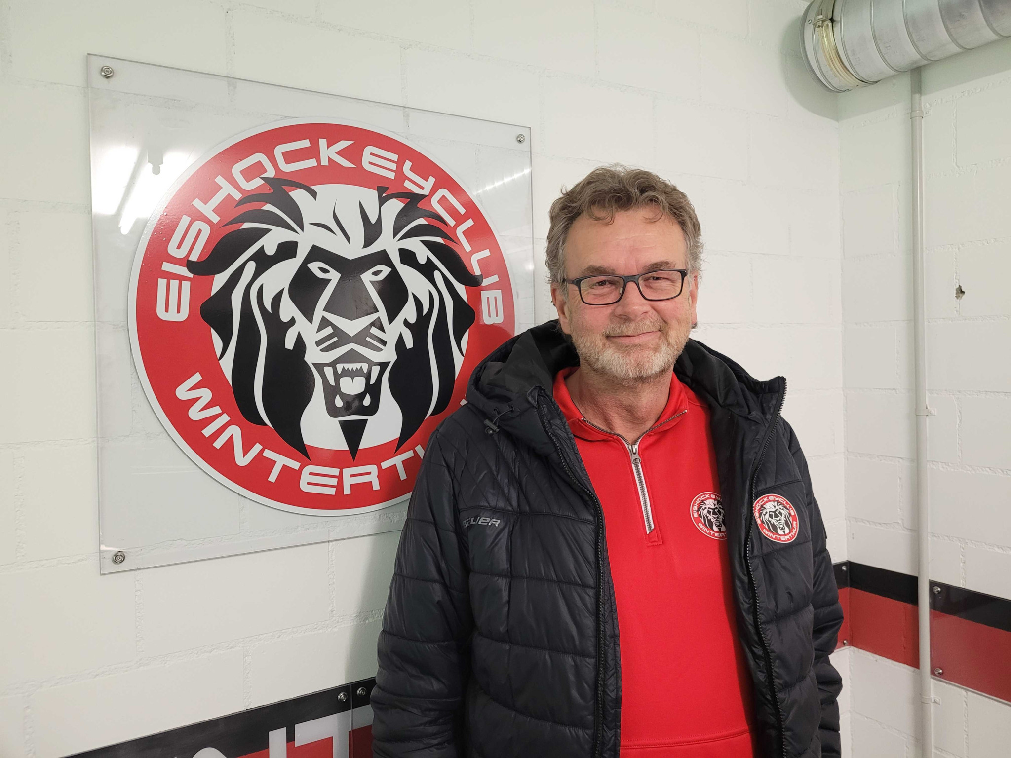 Mann mit Brille steht vor dem Logo des Eishockeyclubs Winterthur, einem roten Löwenkopf auf weissem Grund.