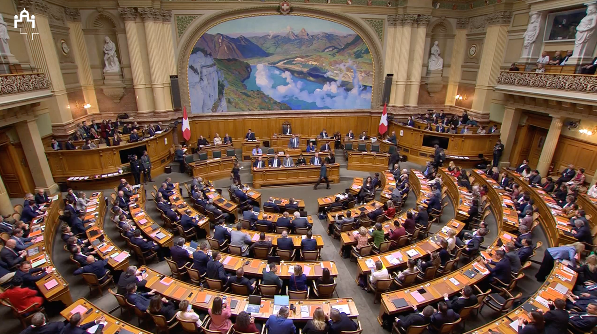 Vue d’une session parlementaire dans un bâtiment historique, avec des députés assis autour de tables disposées en demi-cercle.