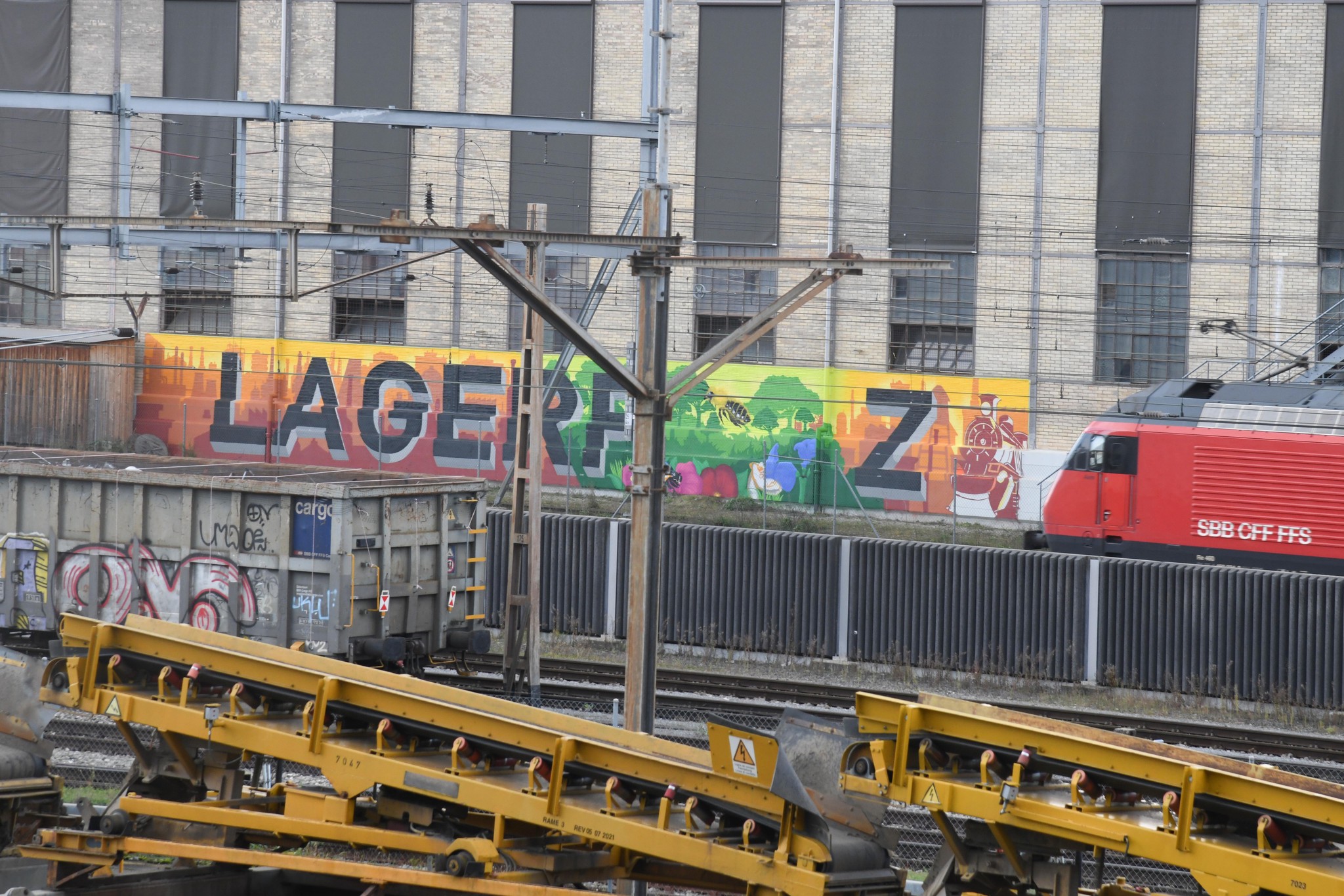 Graffiti mit dem Schriftzug 'Lagerplatz' und leuchtenden Farben an einer Wand in Winterthur, neben Gleisen und einem roten SBB CFF FFS Zug.