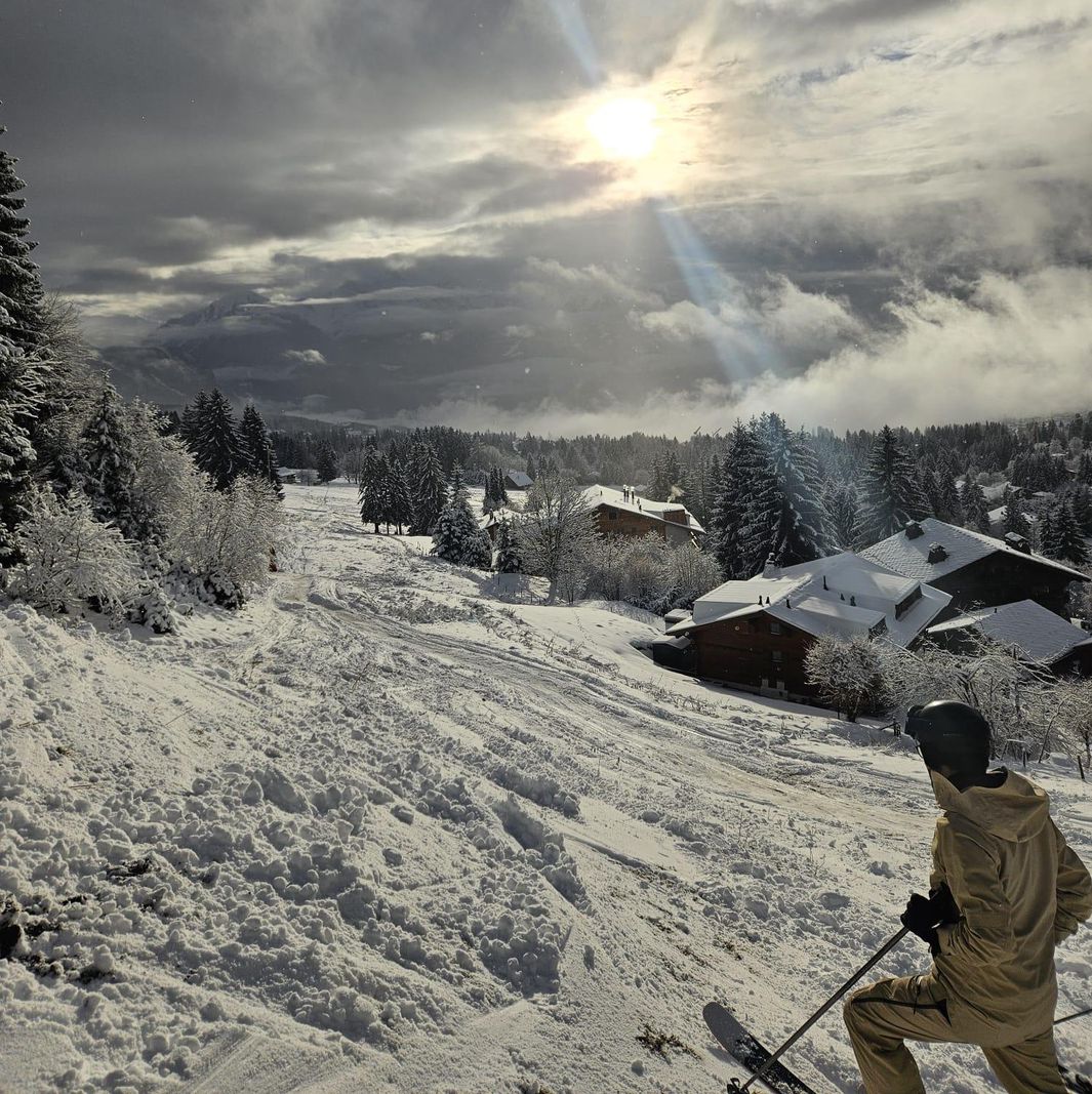 Skieur sur une piste enneigée, entourée de chalets et d’arbres sous un ciel nuageux avec des rayons de soleil perçant.