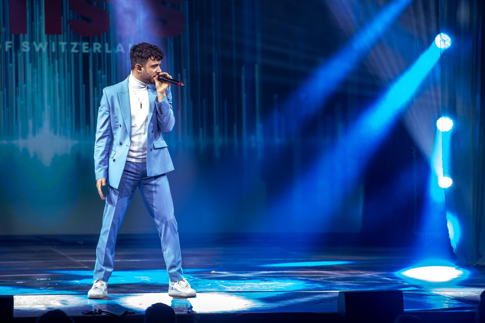 Sänger im hellblauen Anzug performt auf einer Bühne mit blauem Scheinwerferlicht, im Hintergrund Schriftzug «of Switzerland».