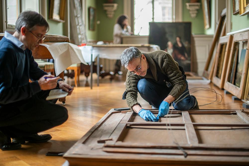 Deux personnes travaillent sur le cadre d’une peinture dans une galerie d’art, l’une en train de prendre des notes et l’autre manipulant les fixations du cadre.