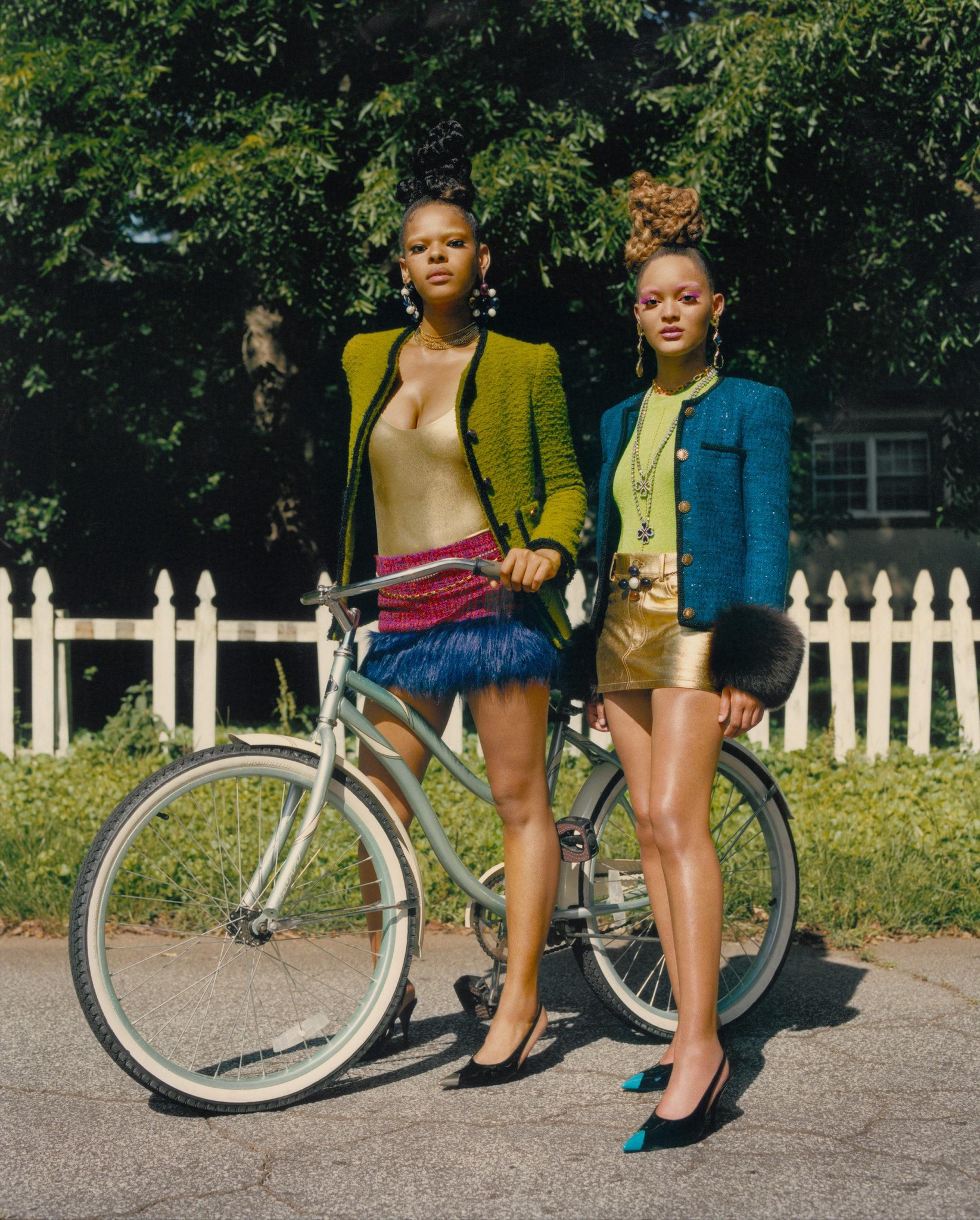 Deux femmes élégamment habillées posent à côté d’un vélo devant une clôture blanche, vêtues de jupes brillantes et de vestes colorées.