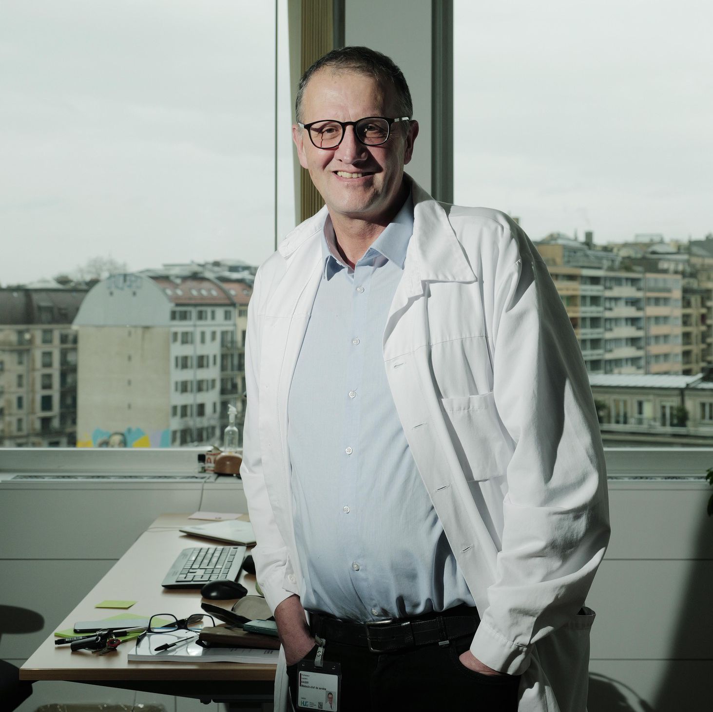 Un professeur en blouse blanche debout dans un bureau aux Hôpitaux Universitaires de Genève, avec vue sur la ville, pour une interview sur cinq ans de la crise Covid.