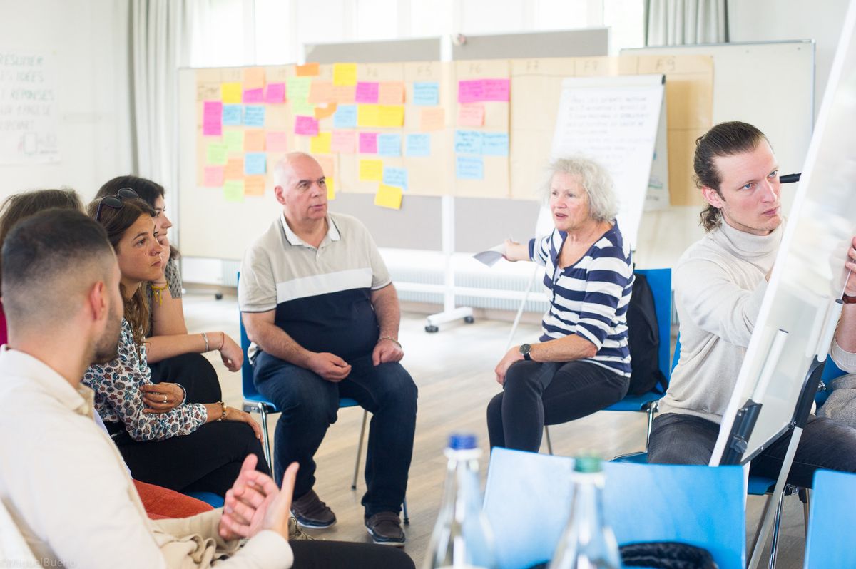Un groupe de personnes assis en cercle lors d'une séance de brainstorming. Des notes colorées sont affichées sur le tableau blanc à l'arrière-plan.