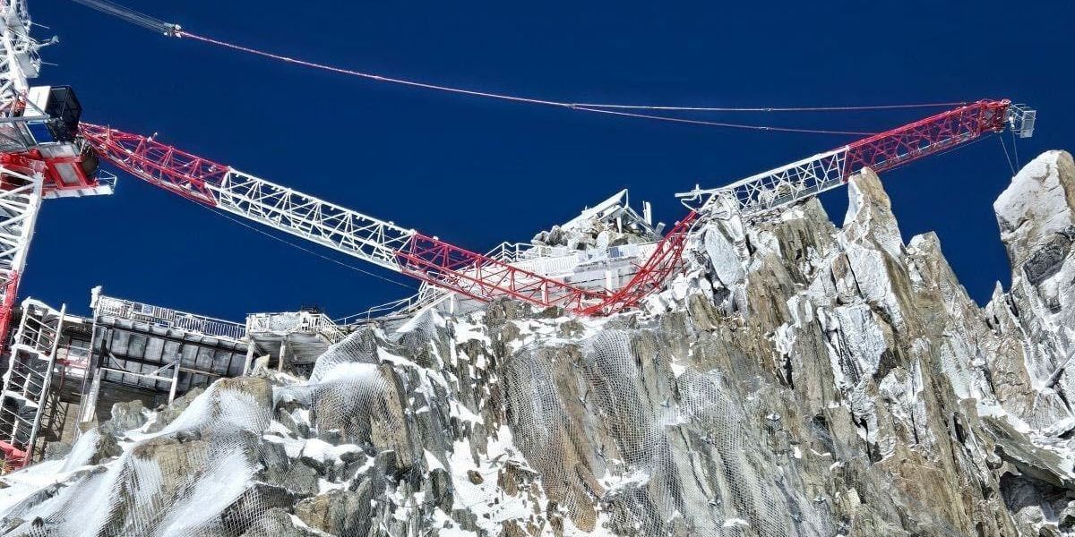 Grue de construction sur un sommet rocheux enneigé sous un ciel bleu vif.