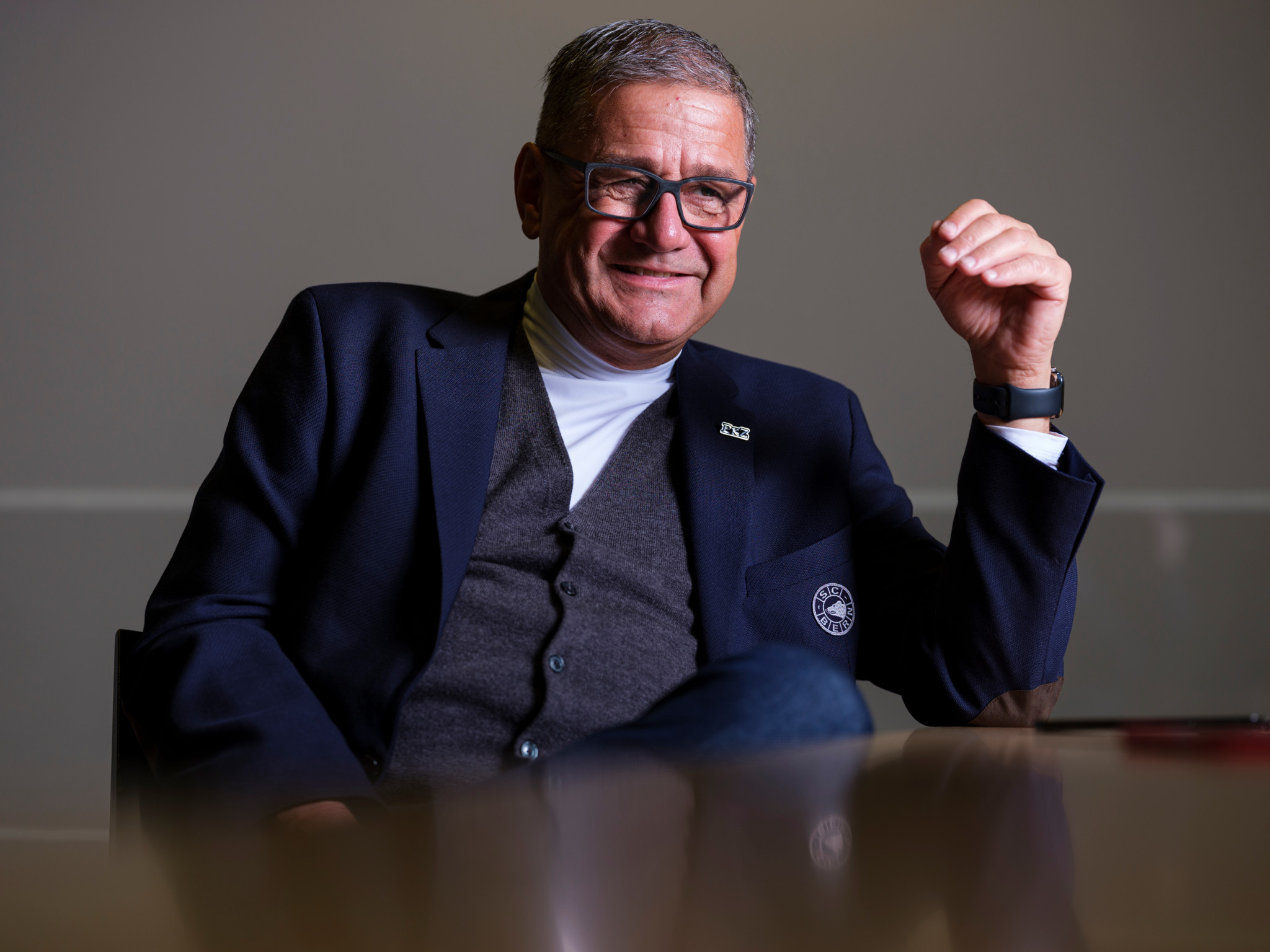SCB-CEO Marc Lüthi sitzt an einem Tisch, lächelt und gestikuliert mit der Hand, gekleidet in Blazer und Cardigan.
