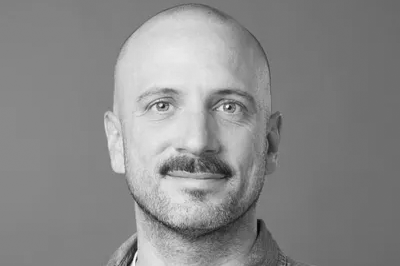 Portrait en noir et blanc d’un homme chauve avec une moustache, portant une chemise en jean.