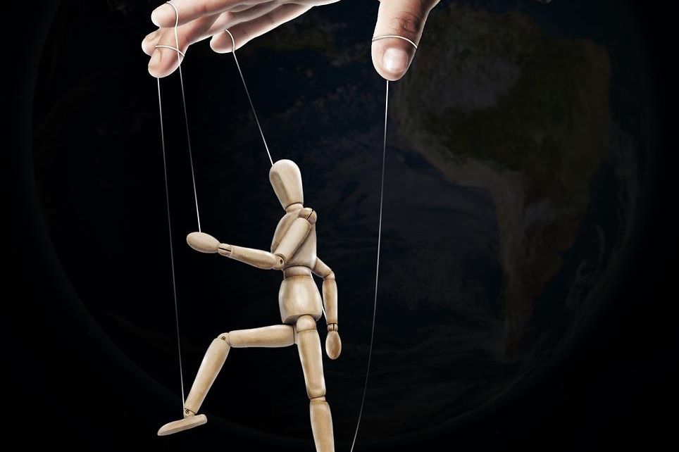 Une main manipule une marionnette en bois avec des fils, symbolisant le contrôle ou la manipulation.