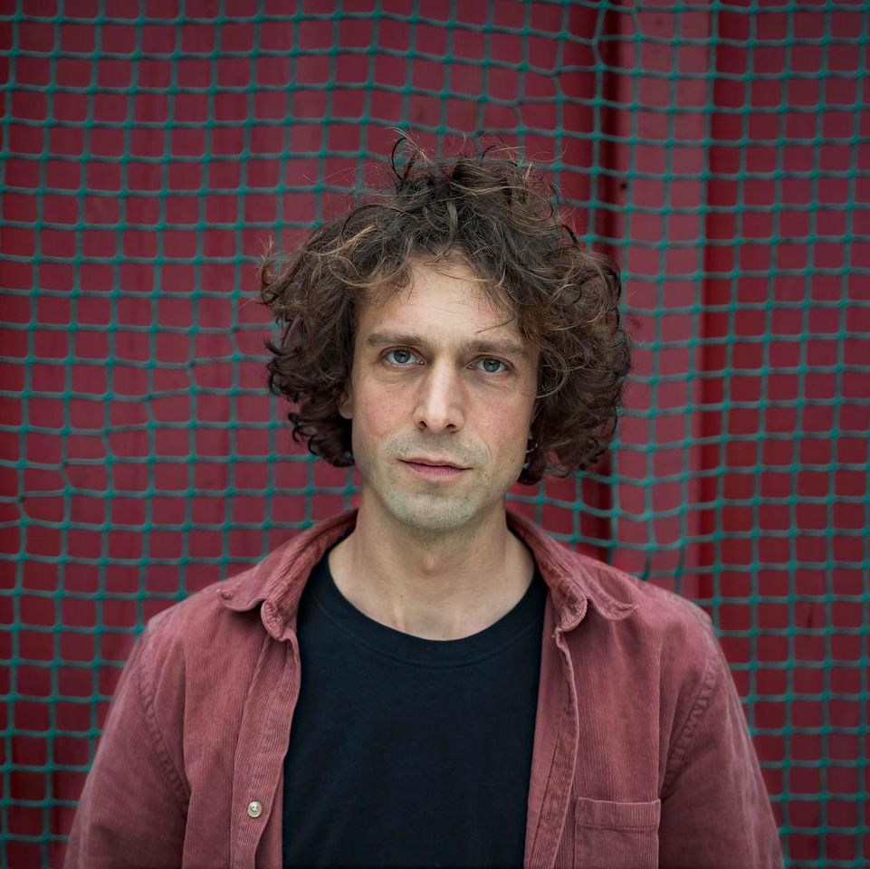 Un homme aux cheveux bouclés pose devant un filet avec un fond rouge.