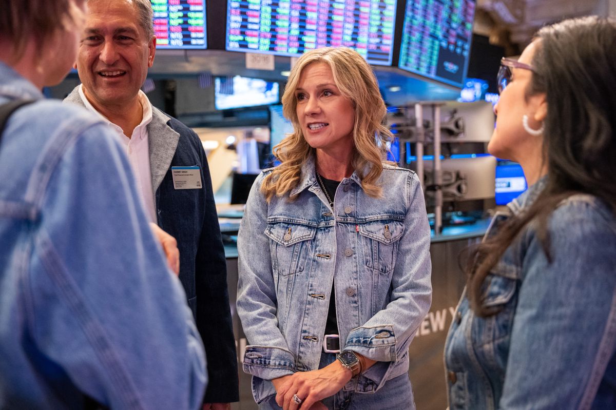 Michelle Gass, PDG de Levi Strauss & Co., se tient sur le parquet de la Bourse de New York pendant un événement de cotation de Levi's, discutant avec des collègues. Plusieurs écrans affichant des données boursières sont visibles en arrière-plan.