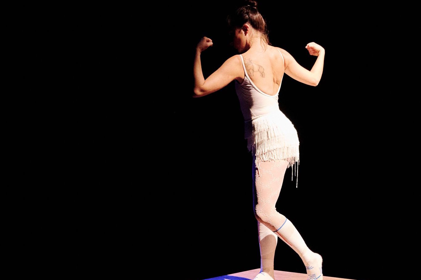 Danseuse en justaucorps blanc posant avec les bras fléchis sur une boîte en bois, éclairée par une lumière dramatique dans un espace sombre.