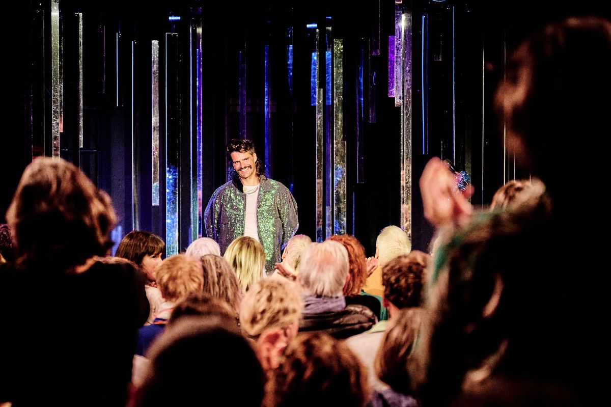 Un homme souriant sur une scène applaudit par une foule d'un public diversifié pendant un spectacle.