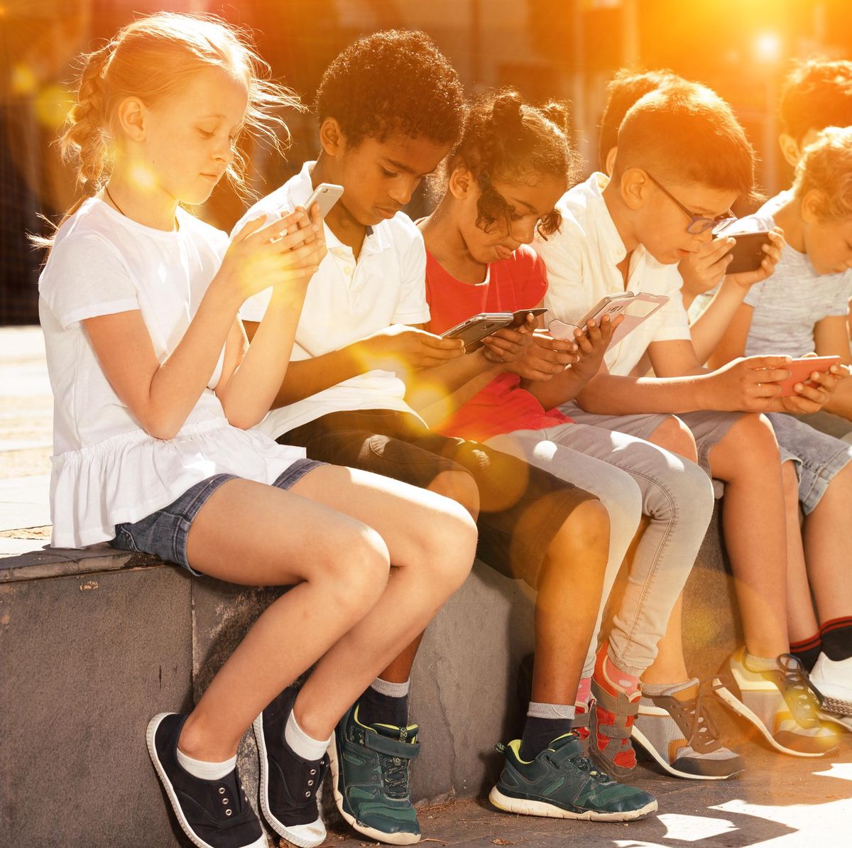 Des enfants jouent sur des smartphones dans un parc, assis en ligne sur un muret.