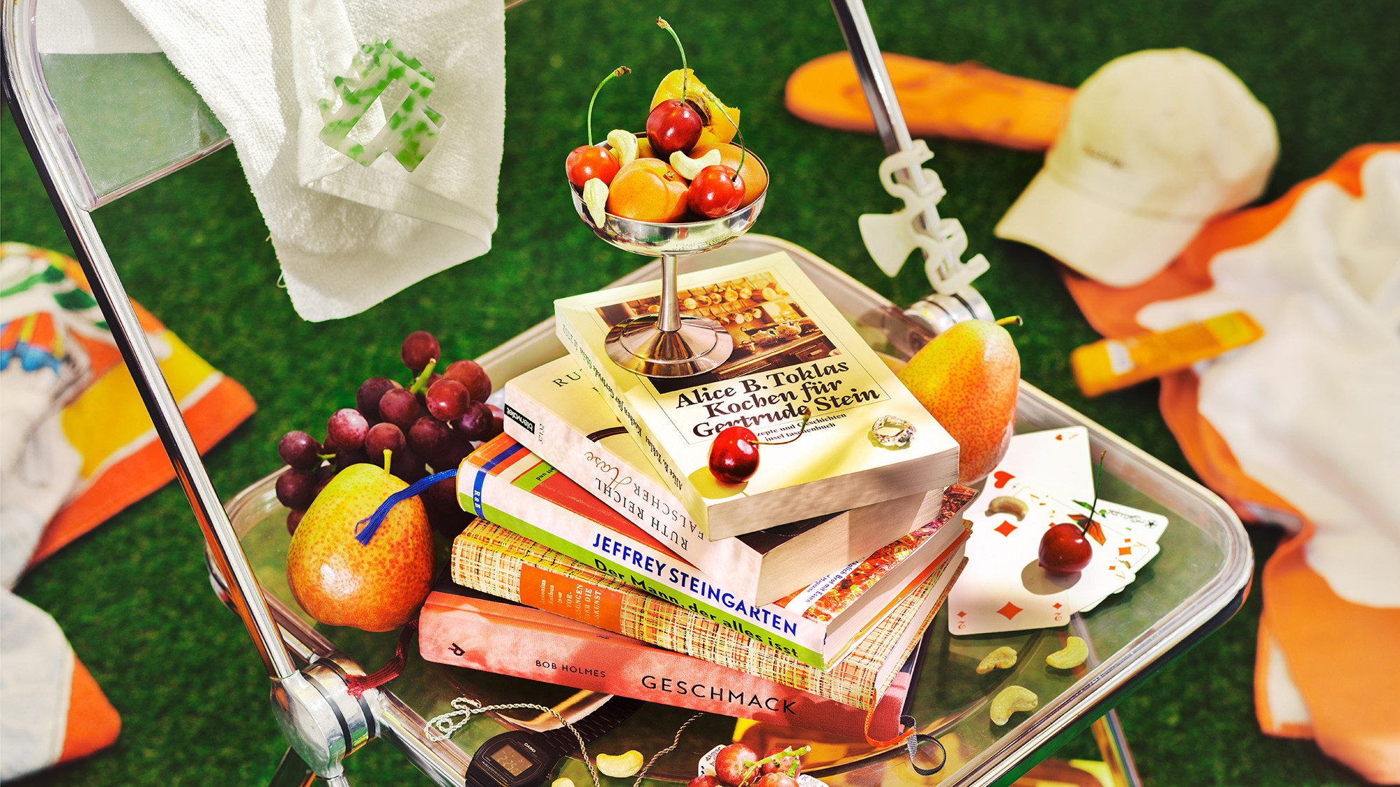 Stapel bunter Kochbücher, darunter Werke von Jeffrey Steingarten und Alice B. Toklas, dekoriert mit frischen Früchten auf einem Servierwagen im Grünen.