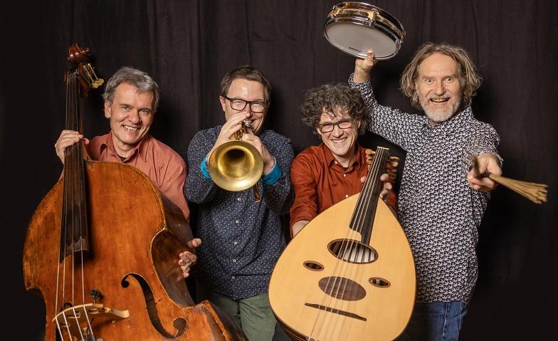 Eine lächelnde Musikgruppe posiert mit Musikinstrumenten: Kontrabass, Trompete, Oud und Tamburin vor einem dunklen Hintergrund.